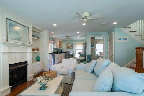 A cozy living room with light blue furniture, a fireplace, and an open layout leading to the kitchen. The space features a ceiling fan, staircase, and various decorative elements.