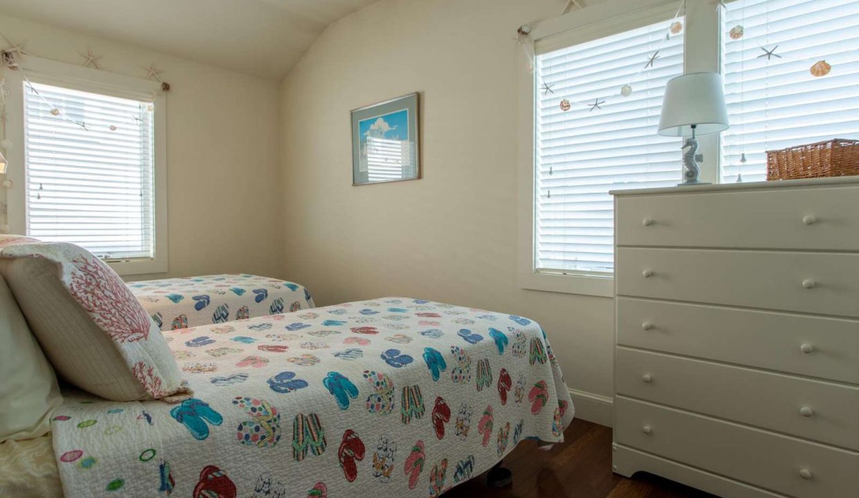 A bedroom with two twin beds featuring colorful quilts, a white dresser with a lamp, and two windows with blinds. A coastal-themed picture hangs on the wall between the windows.