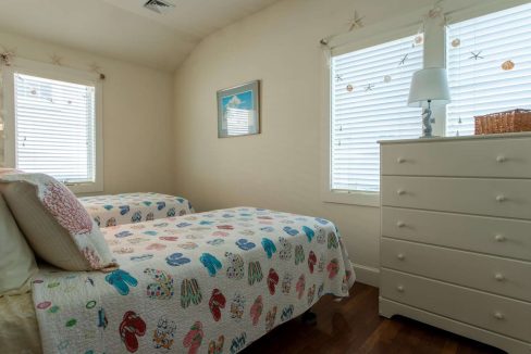 A bedroom with two twin beds featuring colorful quilts, a white dresser with a lamp, and two windows with blinds. A coastal-themed picture hangs on the wall between the windows.