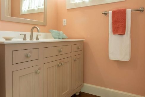 A bathroom with salmon-colored walls, a window with a white curtain, a large mirror, a white vanity with silver knobs, and a towel bar holding two towels: one white and one red.