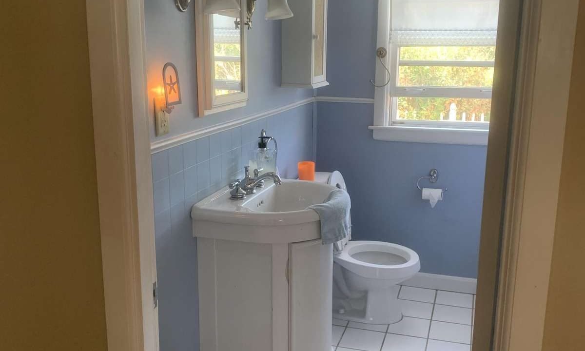 A small bathroom with blue walls, a white sink underneath a mirror, a toilet, and a window with a shade. There is a wall-mounted cabinet and a few toiletries on the sink. A white rug sits on the tiled floor.