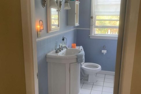 A small bathroom with blue walls, a white sink underneath a mirror, a toilet, and a window with a shade. There is a wall-mounted cabinet and a few toiletries on the sink. A white rug sits on the tiled floor.
