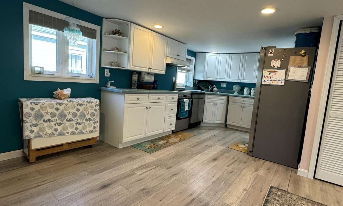 A modern kitchen with white cabinets, stainless steel appliances, a fridge with magnets, teal accent walls, and wooden flooring. A window provides natural light, and a fabric-covered console table is on the left.