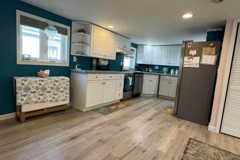 A modern kitchen with white cabinets, stainless steel appliances, a fridge with magnets, teal accent walls, and wooden flooring. A window provides natural light, and a fabric-covered console table is on the left.