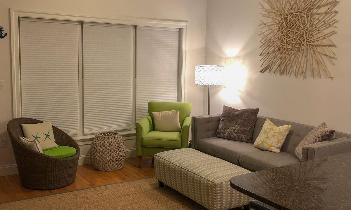 A cozy living room with a sectional sofa, a green armchair, a wicker chair, a cushioned ottoman, a floor lamp, an abstract wall art piece, and a large jute rug on hardwood flooring.
