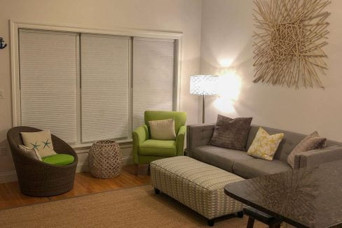 A cozy living room with a sectional sofa, a green armchair, a wicker chair, a cushioned ottoman, a floor lamp, an abstract wall art piece, and a large jute rug on hardwood flooring.
