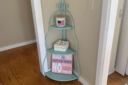 A small teal corner shelf unit with three tiers holds a container, a decorative item featuring the American flag, a small bucket, and a sign that reads 