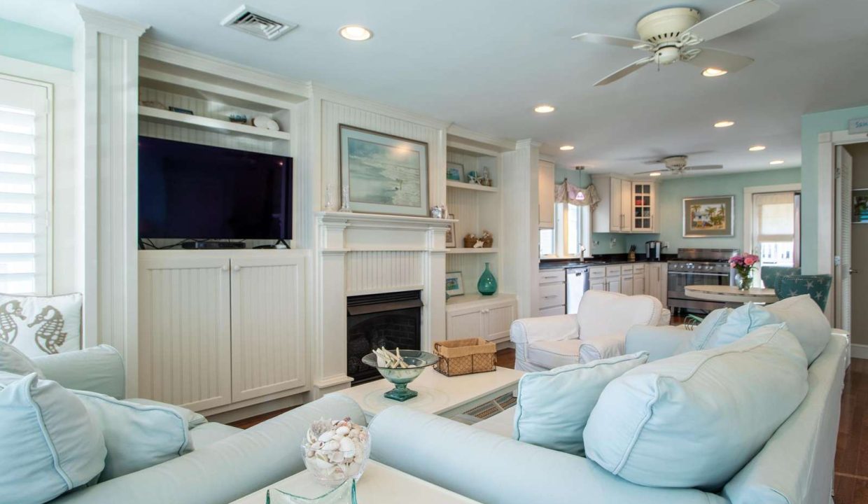 A cozy living room features pastel-colored furniture, a wall-mounted TV above a fireplace, and built-in shelving. The adjacent open kitchen has modern appliances and a ceiling fan overhead.