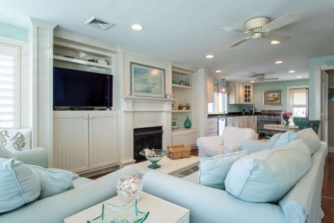 A cozy living room features pastel-colored furniture, a wall-mounted TV above a fireplace, and built-in shelving. The adjacent open kitchen has modern appliances and a ceiling fan overhead.