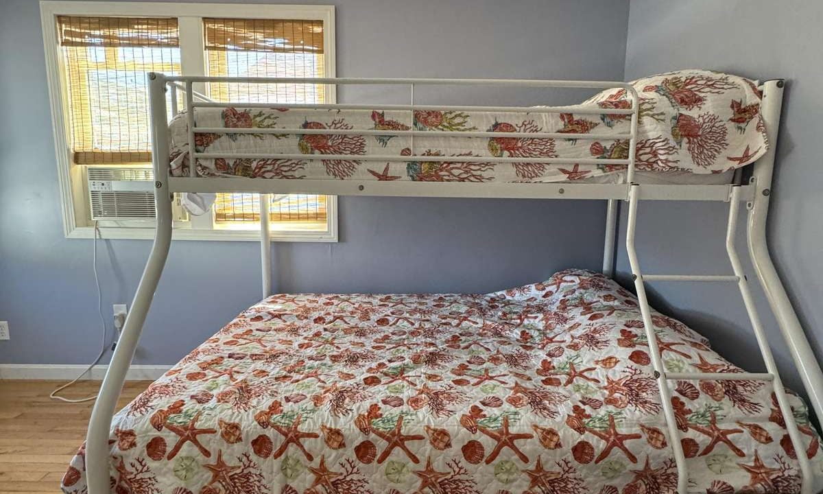 A bedroom with blue walls featuring a metal bunk bed. The top bunk has a single mattress, the bottom bunk has a double mattress. Both mattresses have floral-patterned bedding. A window has bamboo blinds.