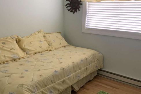 A small bedroom with a bed covered in yellow floral sheets, a window with blinds and a yellow curtain, a sun-shaped wall decoration, and a green rug on a wooden floor.