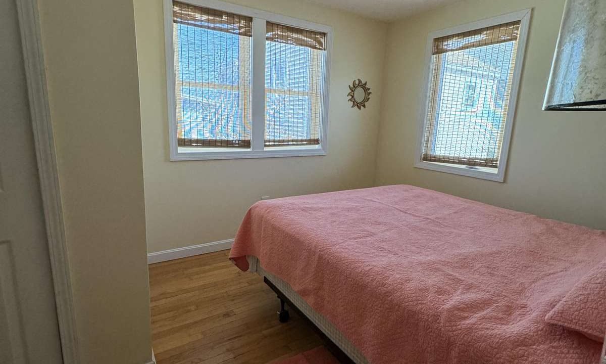 A small bedroom with light-colored walls features a bed with a pink cover and matching rug. The room has wooden floors and three windows with bamboo blinds.