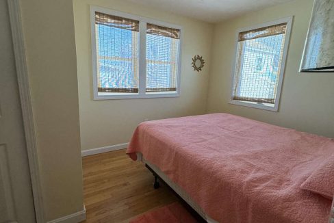 A small bedroom with light-colored walls features a bed with a pink cover and matching rug. The room has wooden floors and three windows with bamboo blinds.