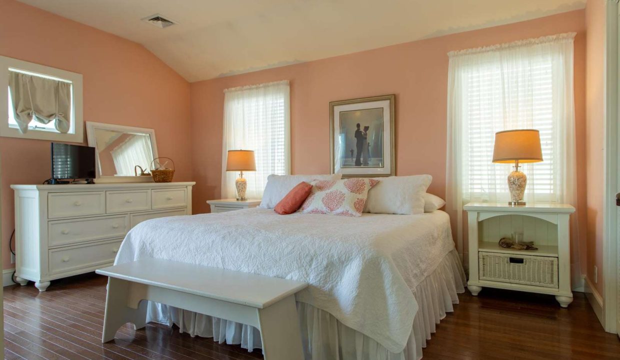 A bedroom with a white king-sized bed, pink walls, two bedside tables with lamps, a dresser with a mirror, a small TV, and a bench at the foot of the bed. There are two windows with sheer curtains.