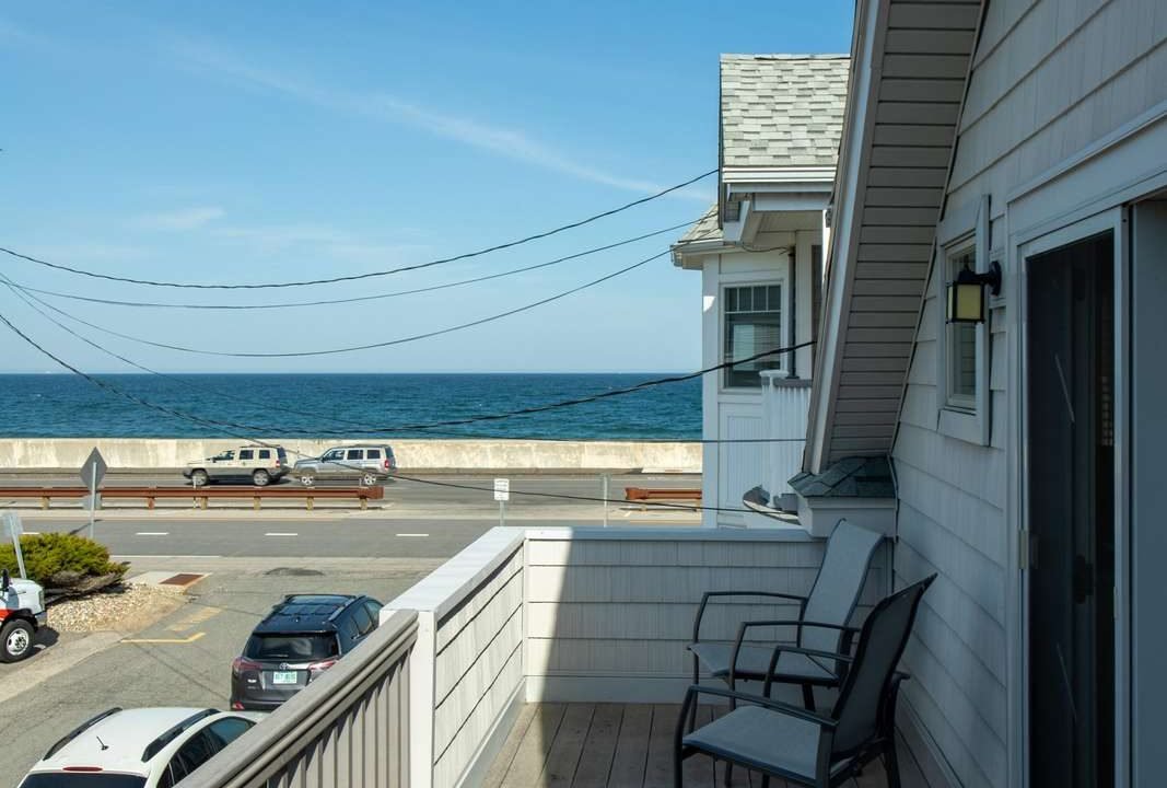 A coastal home's balcony with two chairs overlooks a street and the ocean. A few cars are parked on the street, and the sky is clear and blue.