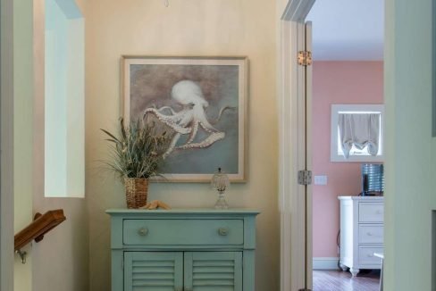 A hallway with wood flooring features a light blue cabinet with a framed octopus artwork above it, a chandelier hanging from the ceiling, and doors leading to adjoining rooms.