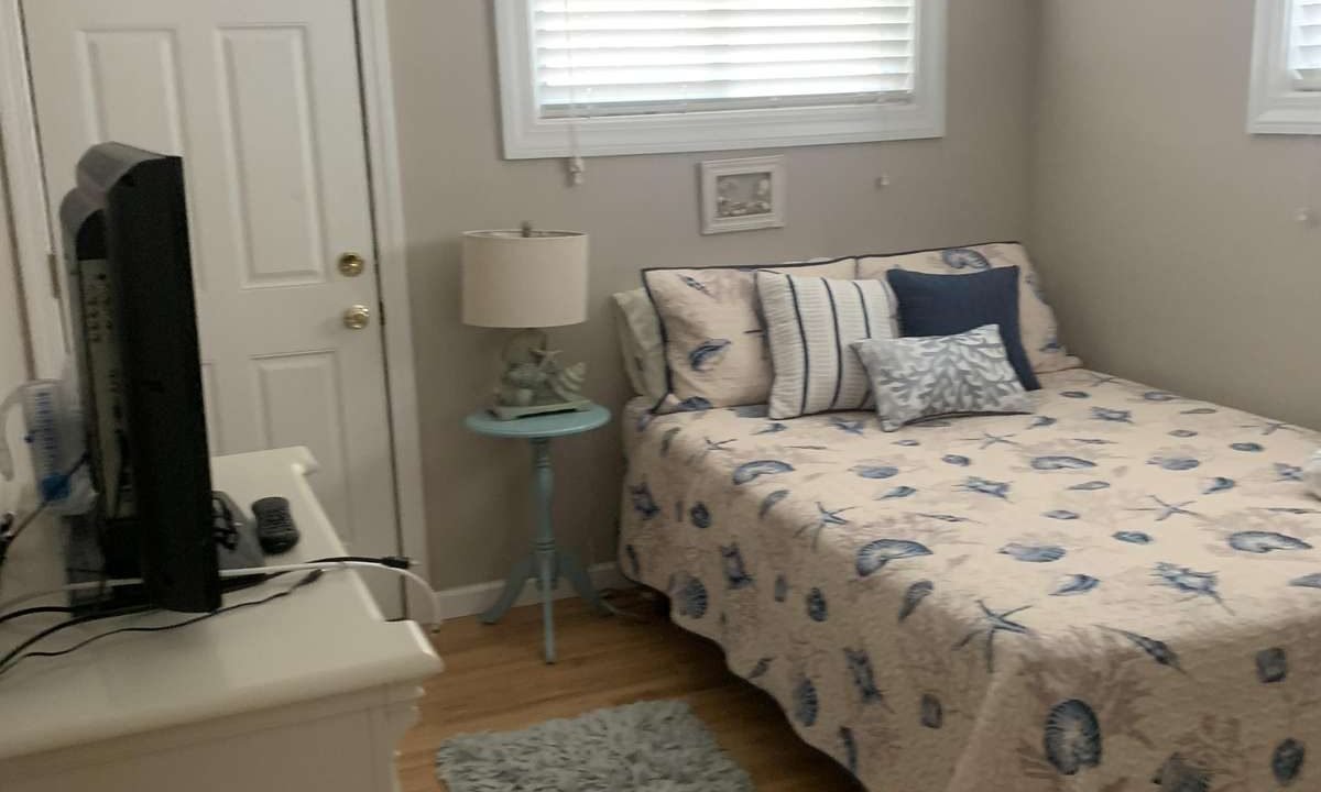Small bedroom with a bed against the right wall, a nightstand with a lamp, a TV on a dresser, and a door to the left. The bed has a nautical-themed quilt and pillows. Light comes from two windows.