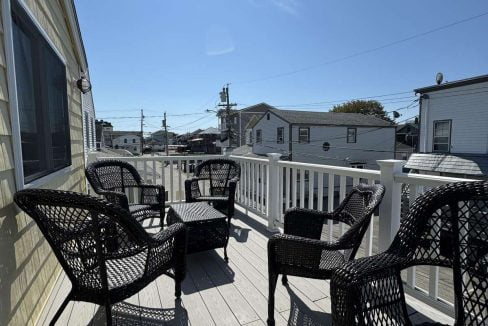 A sunny outdoor deck with five black wicker chairs arranged around a small area, bordered by white wooden railings. Several houses and power lines are visible in the background.