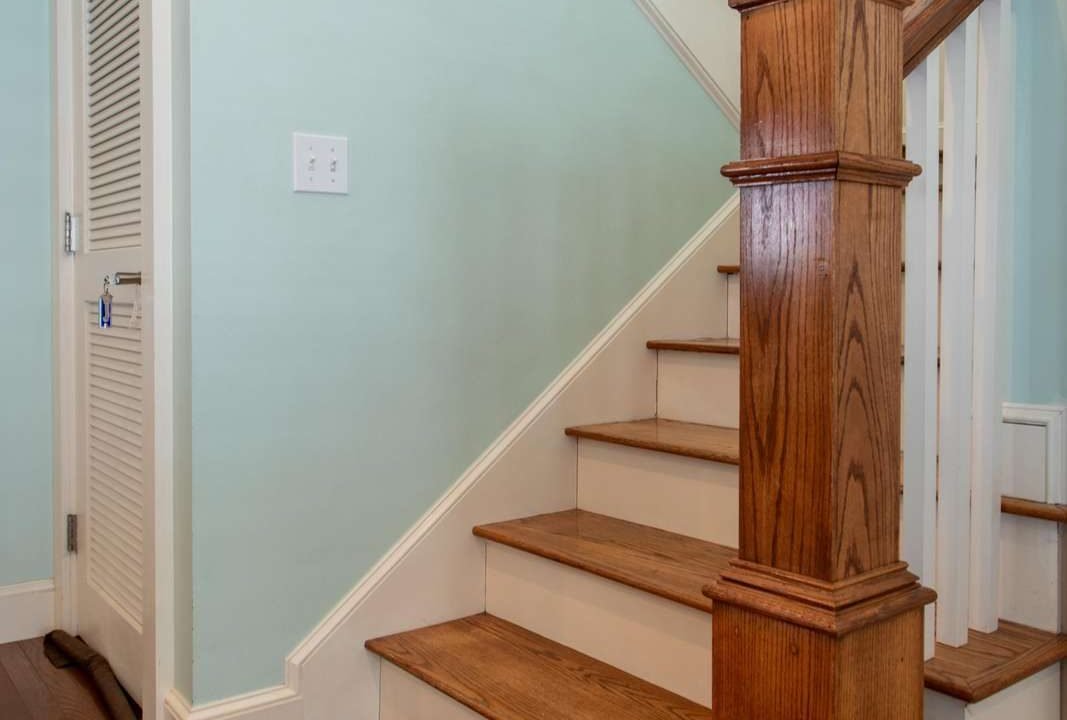 A small staircase with wooden steps and white railing, located next to a white door and framed pictures on a light blue wall.