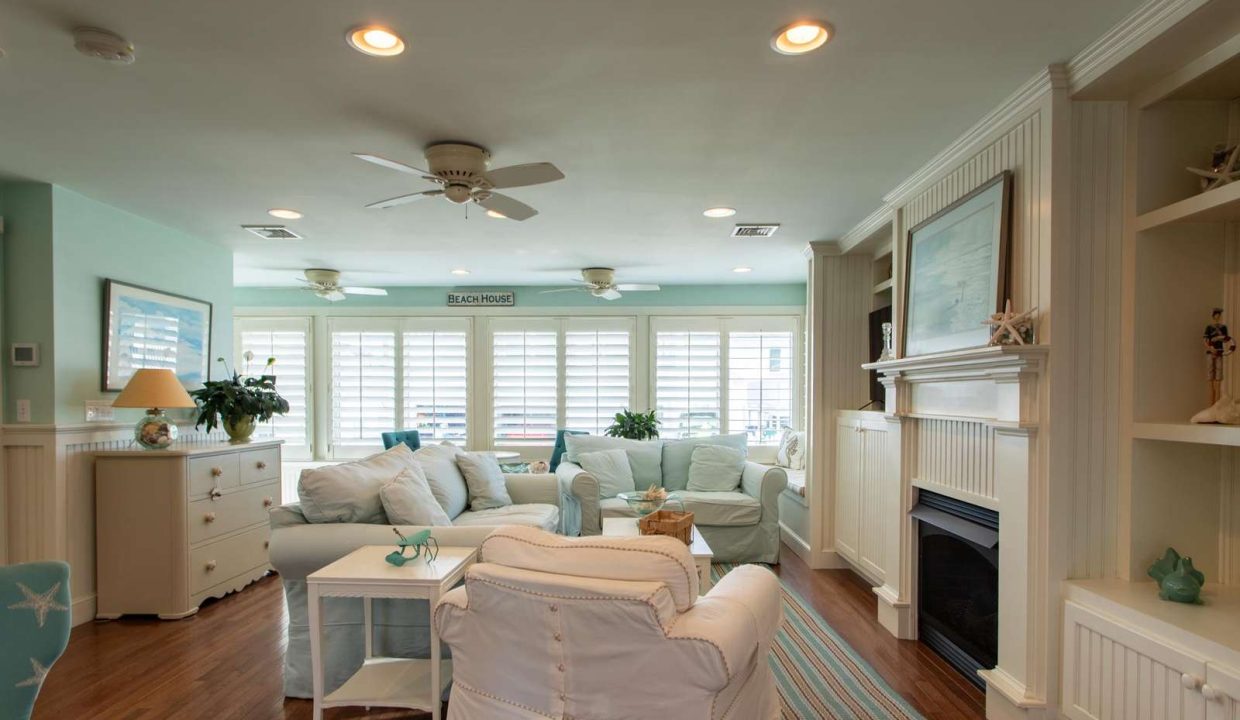 A spacious living room with light blue and white decor, featuring multiple seating options, a fireplace, built-in shelves, and large windows with shutters letting in natural light.