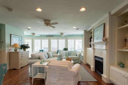 A spacious living room with light blue and white decor, featuring multiple seating options, a fireplace, built-in shelves, and large windows with shutters letting in natural light.