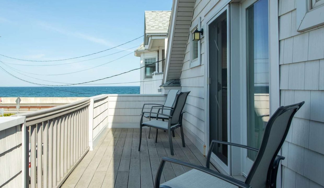 A wooden balcony with two chairs overlooks the ocean. The sky is clear, and nearby buildings and power lines are visible.