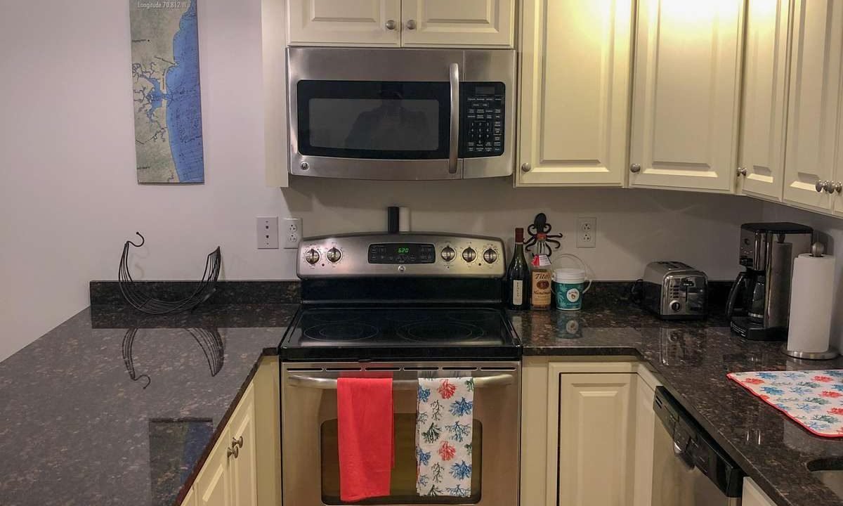 A modern kitchen with white cabinets, stainless steel appliances, and dark granite countertops. Two towels hang on the oven door, and various small appliances sit on the counters.