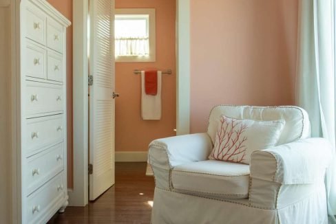 A cozy room with peach-colored walls, a white armchair with a coral-patterned pillow, a white dresser, and an open door leading to a bathroom with matching peach walls and an orange-striped towel.
