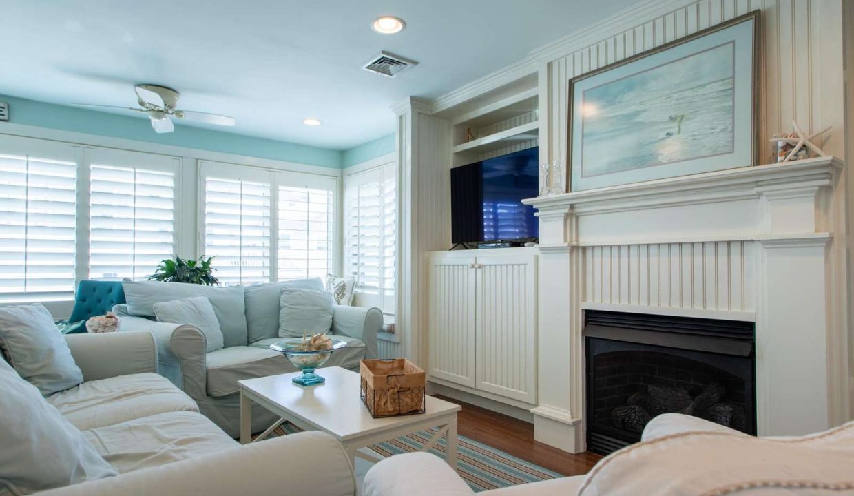 A cozy living room features light-colored furniture, a fireplace with built-in shelves, a television, and large windows with shutters. A ceiling fan and framed art are also visible.