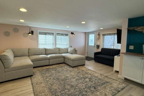 A living room with a large beige sectional sofa, patterned area rug, mounted TV, and light-wood flooring. Natural light filters through windows with blinds.