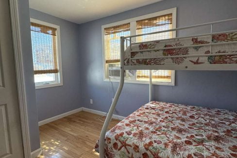 A bedroom with light blue walls, wood flooring, and two windows with bamboo blinds. It has a white metal bunk bed with a seashell-themed bedspread.