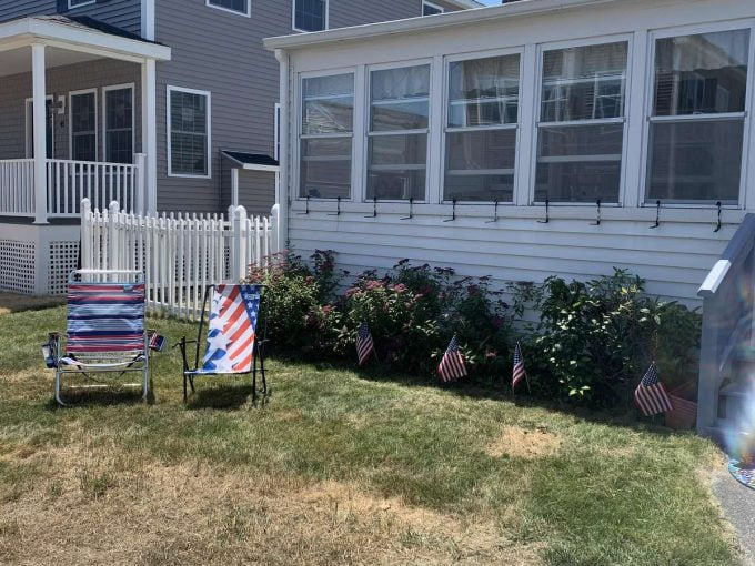 Two lawn chairs face a white house with a lined row of small American flags in the front yard. A neighboring house is visible on the left. The sky is clear and sunny.