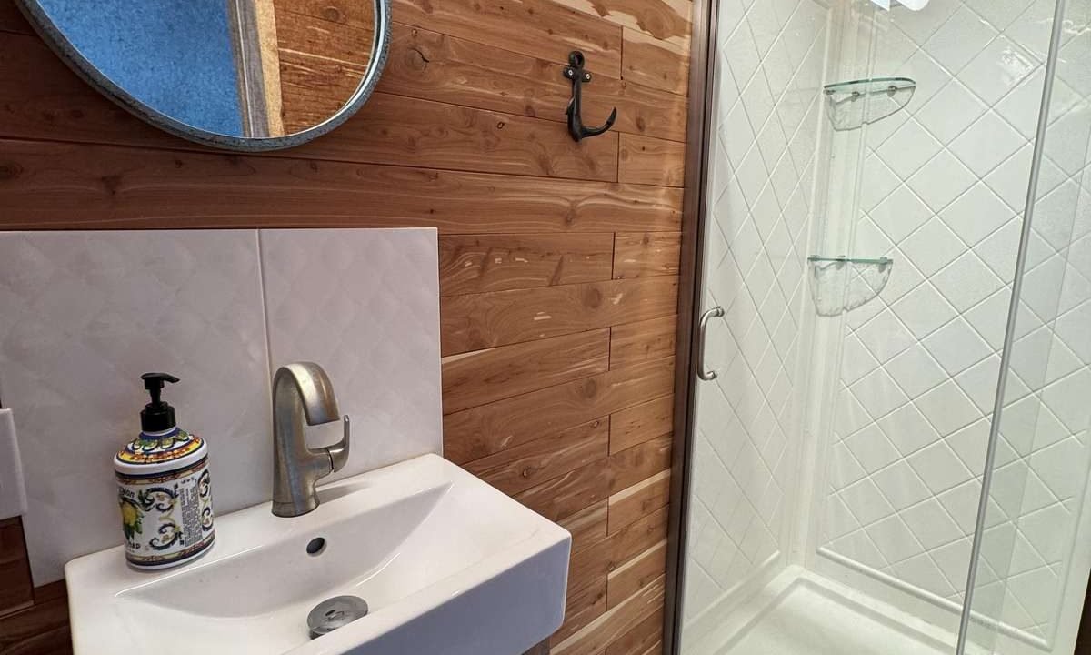 A small bathroom with wood-paneled walls, a round mirror, a sink with a faucet, and a soap dispenser on the left. On the right, there is a glass-enclosed shower with a white tiled interior.