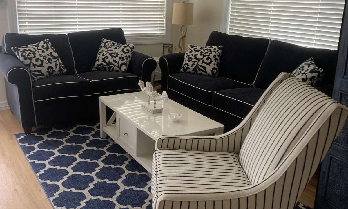 A living room with a navy, white, and beige color scheme featuring a striped armchair, two dark blue sofas, a white coffee table on a patterned blue rug, and blinds on the windows.