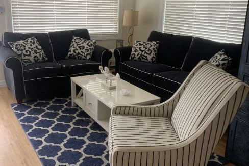 A living room with a navy, white, and beige color scheme featuring a striped armchair, two dark blue sofas, a white coffee table on a patterned blue rug, and blinds on the windows.