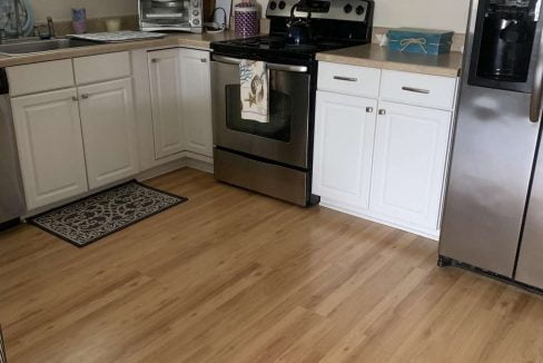 A modern kitchen with white cabinets, stainless steel appliances including a microwave and refrigerator, a black stove, and light wood flooring. A small rug is on the floor near the stove.