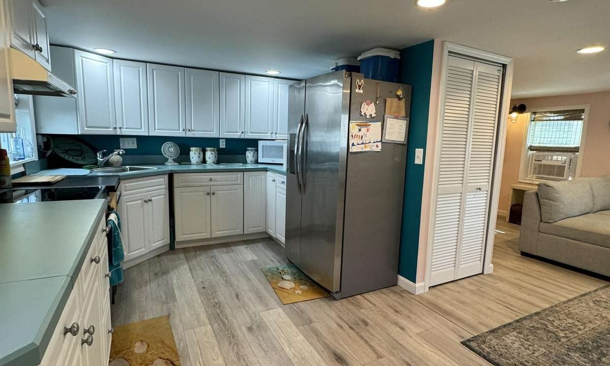 A modern kitchen with white cabinets, a stainless steel refrigerator, and a light gray wooden floor. A starfish-themed rug lies in front of the sink. The open layout reveals a glimpse of a living area.