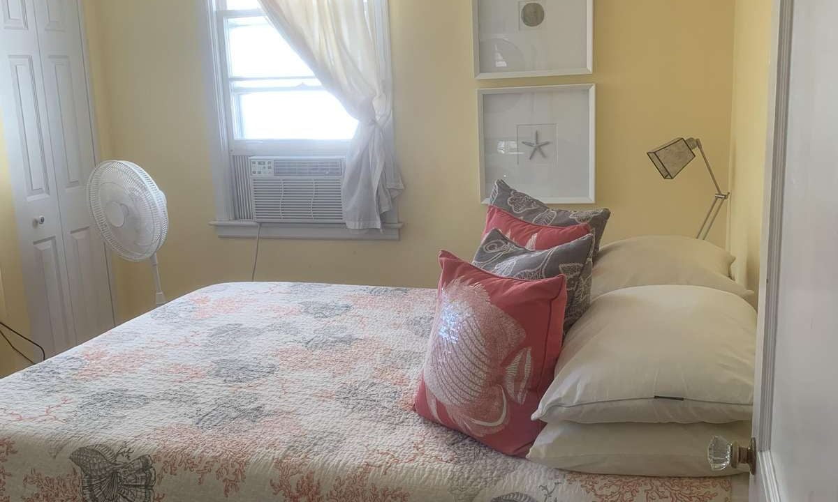 A bedroom with a neatly made bed covered in a coral and seashell-themed quilt, adorned with matching pillows. A window with a sheer curtain, a fan, and minimalistic wall decor can be seen.