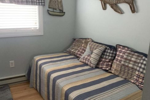 A small bedroom with a twin bed featuring a striped comforter and multiple plaid pillows. The walls are light blue, and fish-shaped wooden wall decor is present above the bed. A window with a plaid curtain.