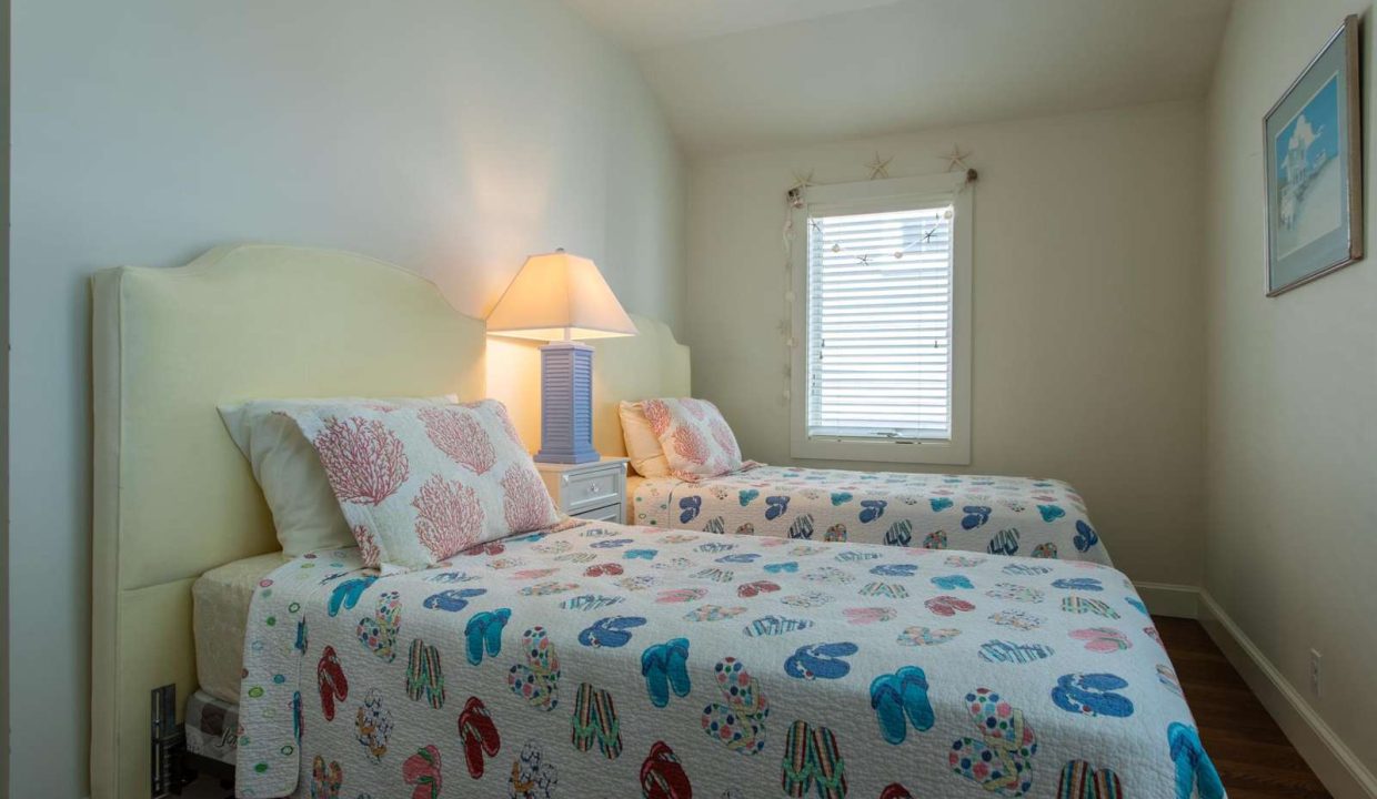 A bedroom with two twin beds covered in colorful patterned quilts, a nightstand with a lamp between them, and a window with blinds.