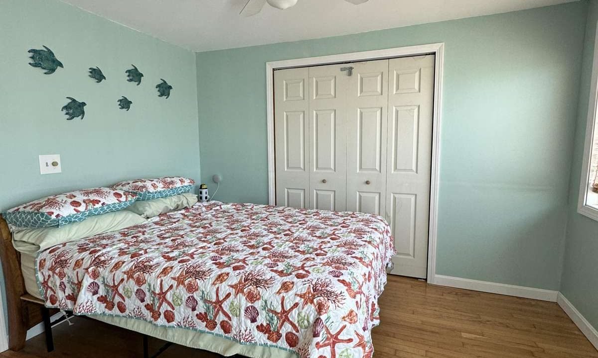 A bedroom with light blue-green walls, a ceiling fan, a double bed with a floral quilt, fish wall decor, a blue rug, and a white double door closet.