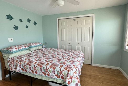 A bedroom with light blue-green walls, a ceiling fan, a double bed with a floral quilt, fish wall decor, a blue rug, and a white double door closet.