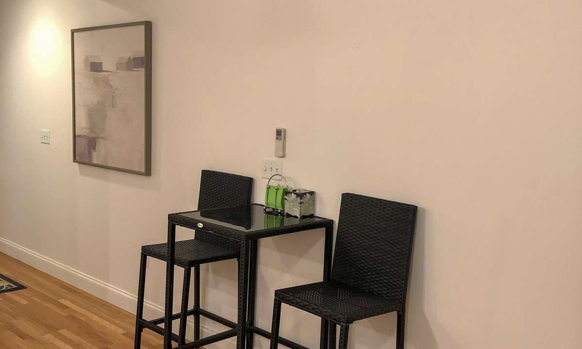 A small table with two tall chairs is set against a plain wall with a framed abstract painting and an air conditioning unit above. The floor is wooden.