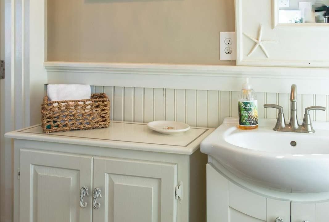 A tidy bathroom features a white sink with a soap dispenser beside a cabinet topped with a basket of towels. A framed picture hangs on the beige wall above, reflecting in a mirror.