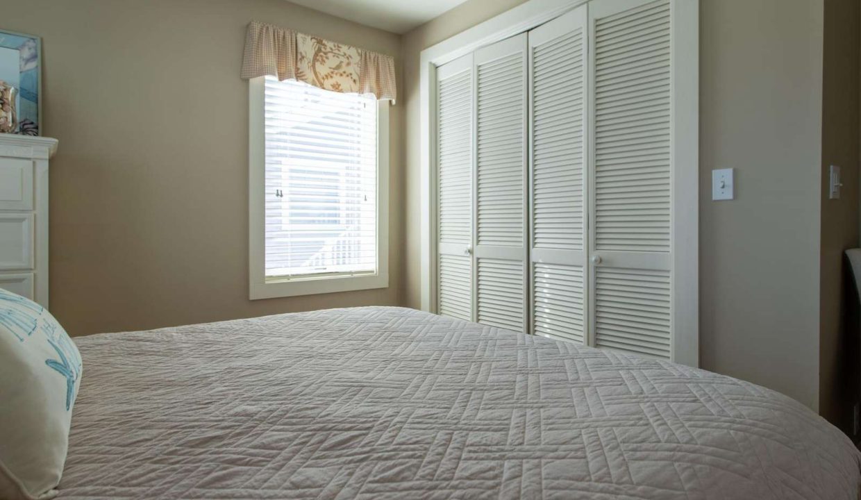 A bedroom with a quilted bedspread, a window with blinds and curtains, and a closed closet with louvered doors.