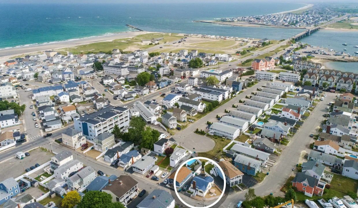 Aerial view of a coastal town with numerous houses, roads, and a visible shoreline. A white circle highlights a specific property in the lower right area of the image.
