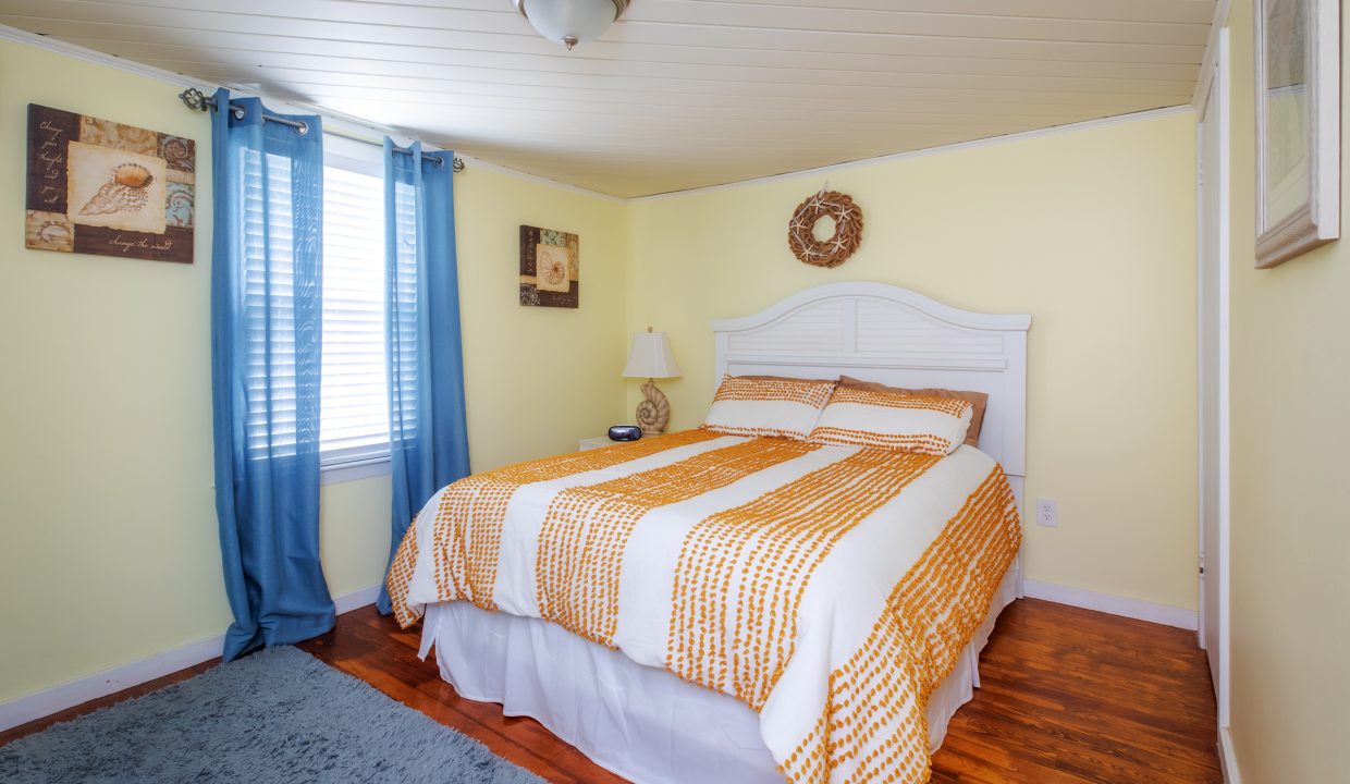 A cozy bedroom with light yellow walls, a white bed with orange and white bedding, blue curtains, and a window on the left. There are two artworks on the walls and a blue rug on the wooden floor.