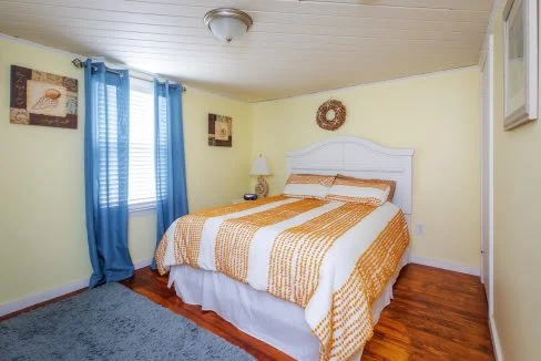 A cozy bedroom with light yellow walls, a white bed with orange and white bedding, blue curtains, and a window on the left. There are two artworks on the walls and a blue rug on the wooden floor.