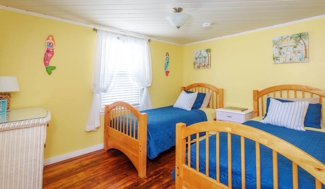 A brightly lit bedroom with yellow walls, two wooden twin beds with blue bedding, a wicker dresser, a white nightstand, and wall decorations. A window with white curtains is centered between the beds.