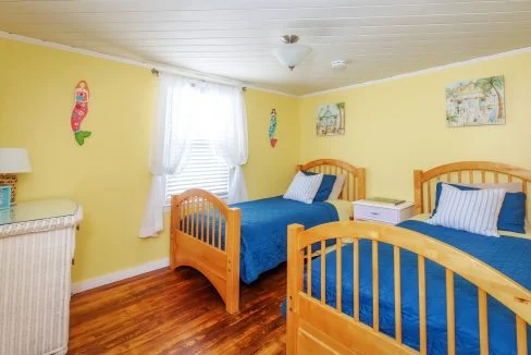 A brightly lit bedroom with yellow walls, two wooden twin beds with blue bedding, a wicker dresser, a white nightstand, and wall decorations. A window with white curtains is centered between the beds.